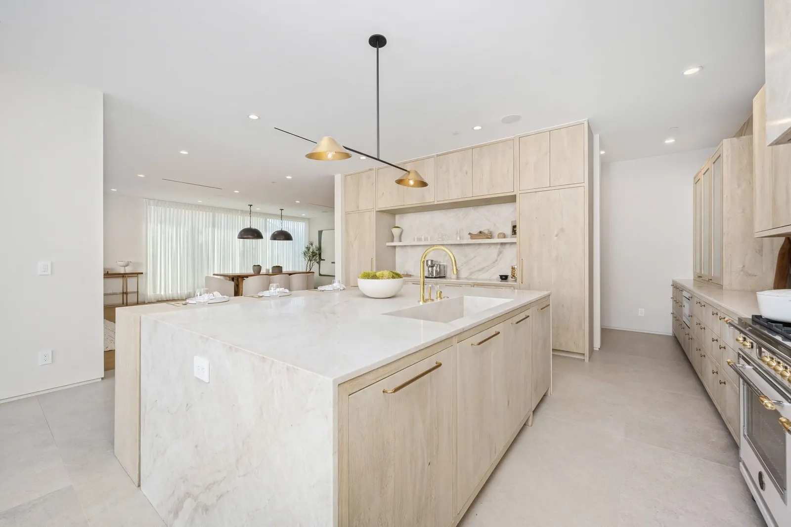 Bright modern kitchen with a large marble island, light wood cabinets, and gold faucet; dining area visible in the background.