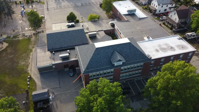 Aerial view of brick building with dark gray roof, trees, and paved areas.