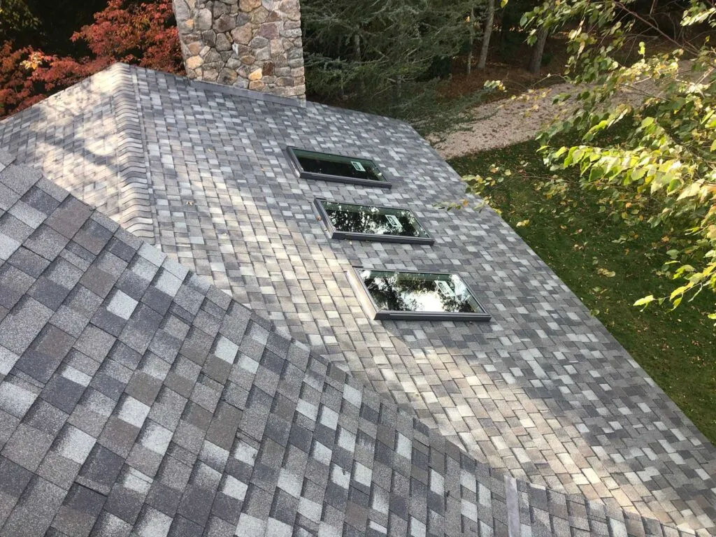 A steep, grey shingled roof features three large rectangular skylights lined up in a row. A stone chimney is visible on the left. The surrounding area has green trees and a small gravel path. The shingle pattern is a mix of light and dark grey tones.