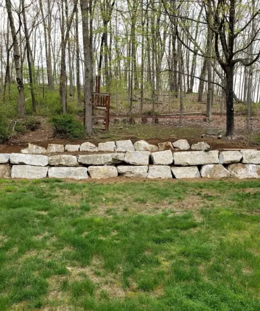 Stone retaining wall beside grassy yard, with wooded area and rusty gate in the background.