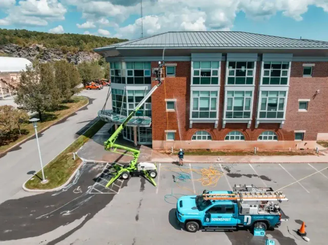 Workers using a green lift to clean the windows of a large brick building while a blue Downspout service truck is parked nearby in an empty parking lot.