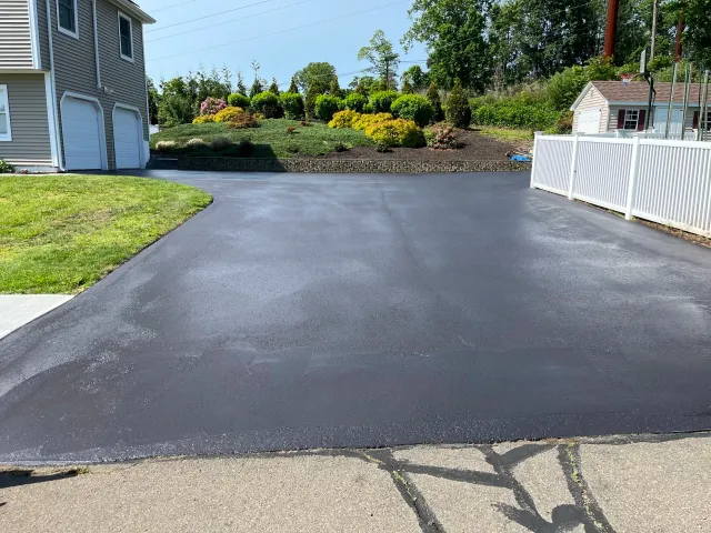 Freshly paved black asphalt driveway adjacent to a grassy lawn, white fence, and landscaped garden with bushes and trees.