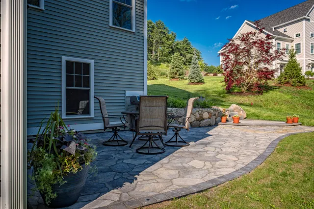 Outdoor patio area with stone flooring, a table with chairs, potted plants, and a landscaped yard with trees.