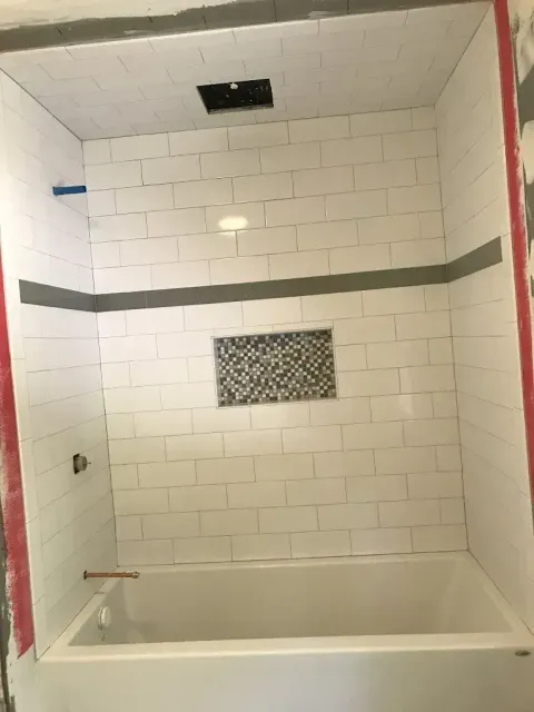 White tiled bathtub shower alcove with a small mosaic accent and unfinished trim.