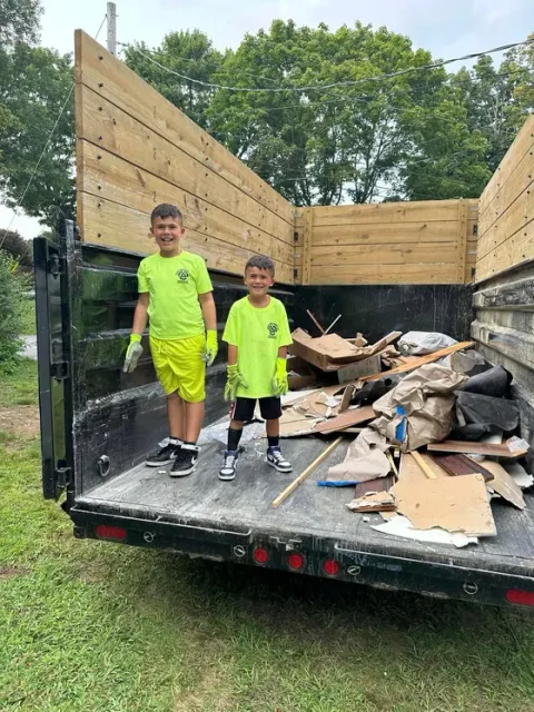Two young boys are standing in the back of a dumpster filled with cardboard.