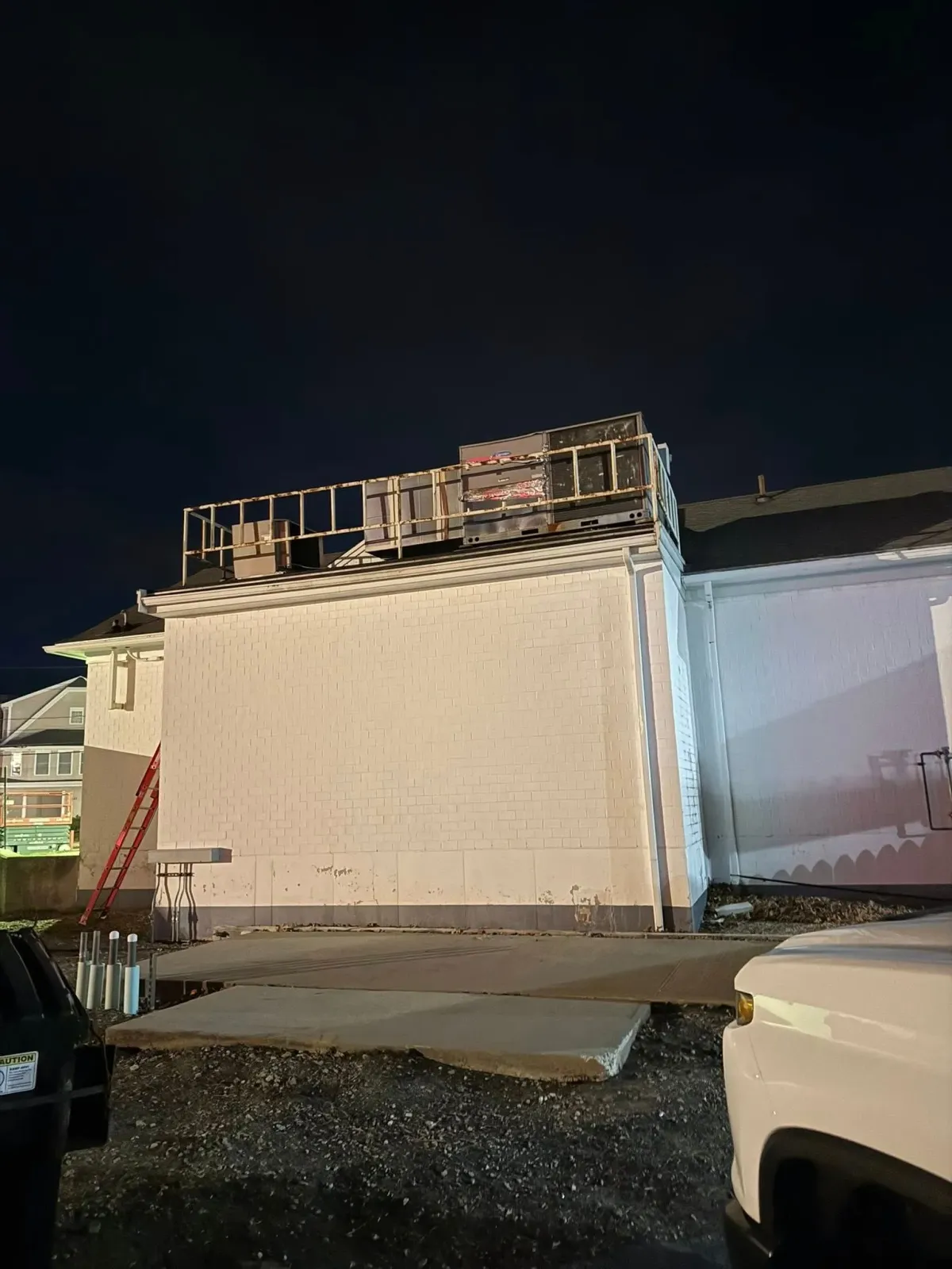 A construction site at night showing a building's white exterior wall with exposed framing on the roof and a red ladder.