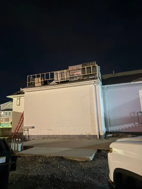 A construction site at night showing a building's white exterior wall with exposed framing on the roof and a red ladder.