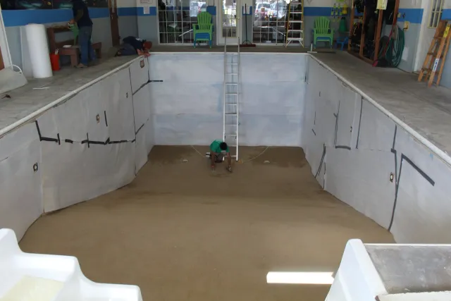 An indoor swimming pool under construction, with a worker preparing the bottom surface and another worker on the side. The pool is empty with a ladder in the center, and walls lined with insulation material. There are chairs and tools stored along th