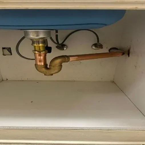 A kitchen sink cabinet with exposed plumbing, showing a copper pipe connected in an unusual L-shaped configuration, lacking a traditional P-trap beneath the sink.
