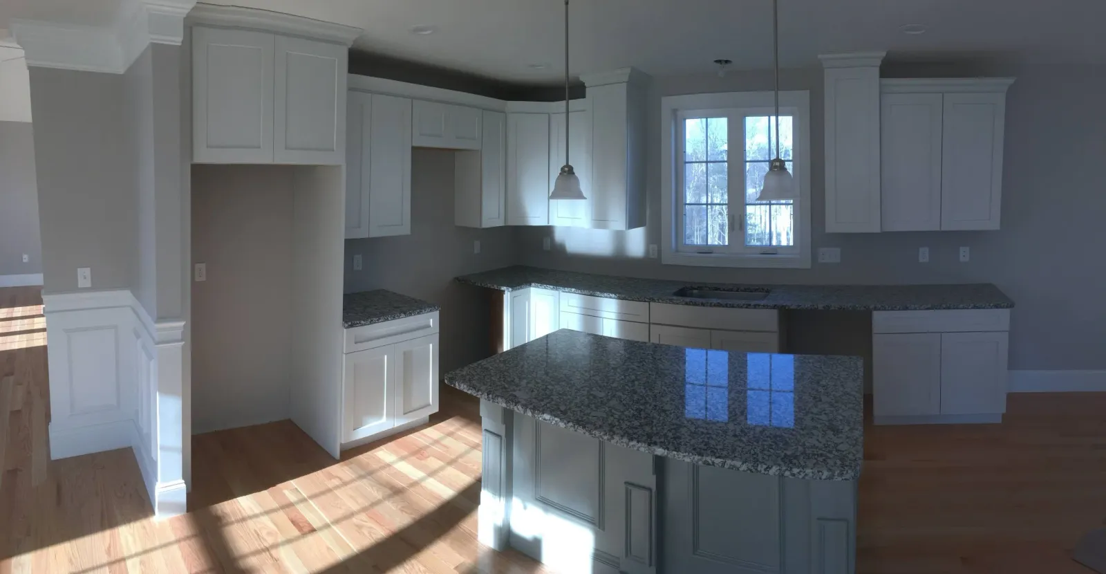 Empty kitchen with white cabinets, granite countertops, wooden floor, large window, pendant lights, and a central island.