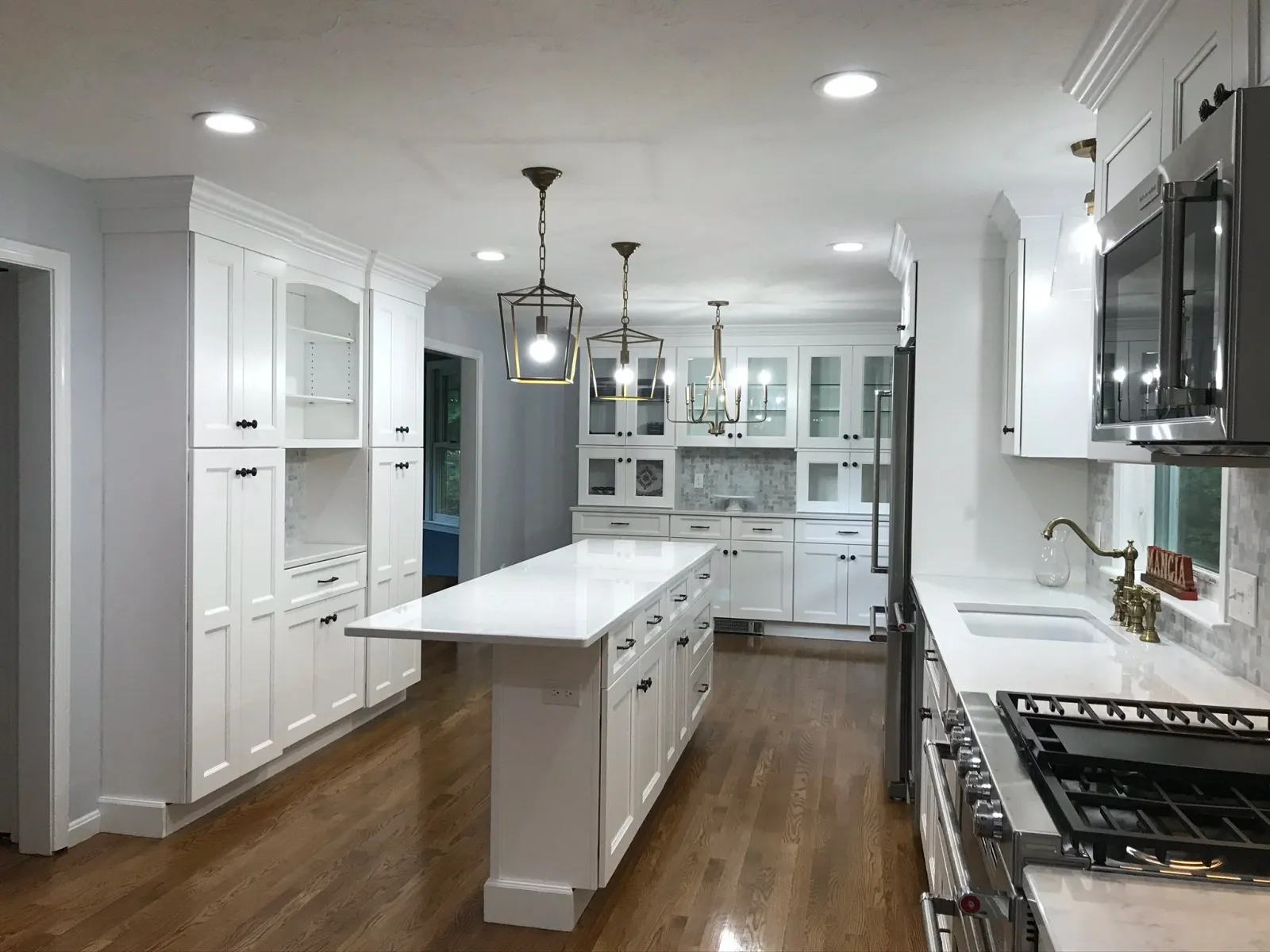 Modern white kitchen with central island, stainless steel appliances, white cabinetry, hardwood floors, and decorative pendant and chandelier lighting.