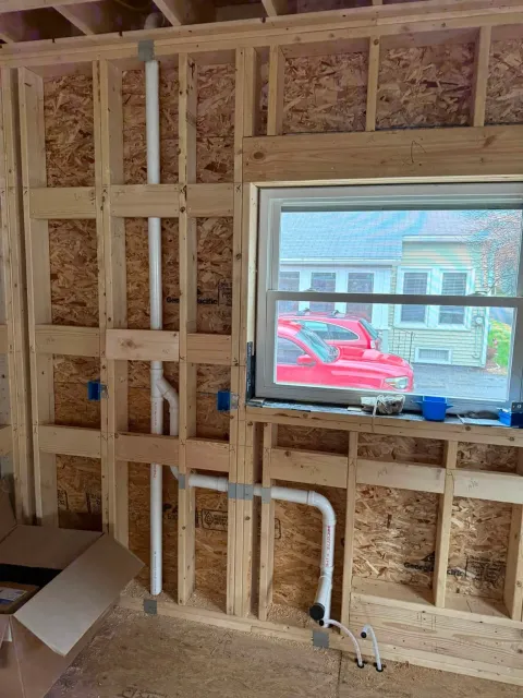 Interior wall framing under construction with a window opening and visible pipes, insulation, and electrical wiring.