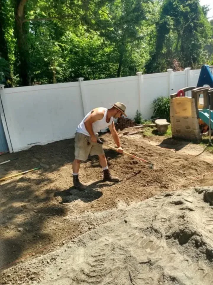 Person raking soil in a backyard with a white fence and playhouse in the background.