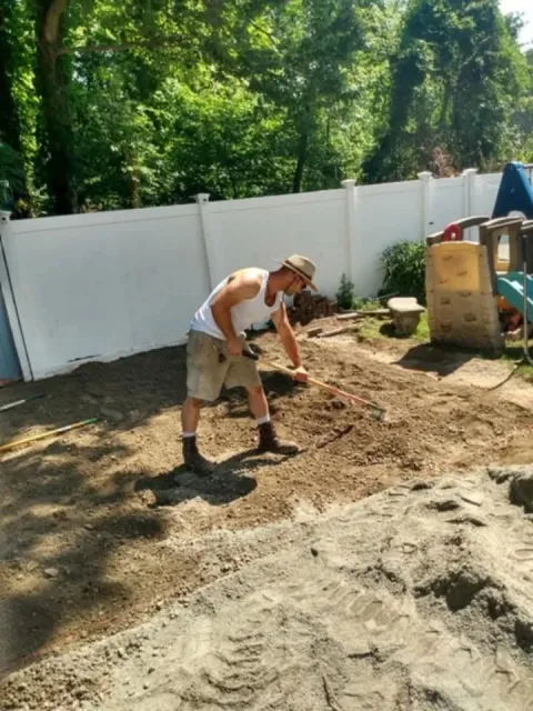 Person raking soil in a backyard with a white fence and playhouse in the background.