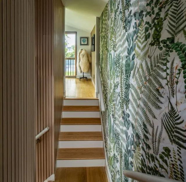 Staircase leading to a small landing with a door and window, decorated with a fur coat on a stand, framed wall art, and wallpaper with a leafy green pattern.