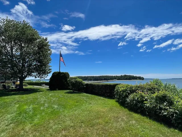 Lawn with a large tree and an American flag on a pole, overlooking a body of water with a distant island and partly cloudy blue sky.