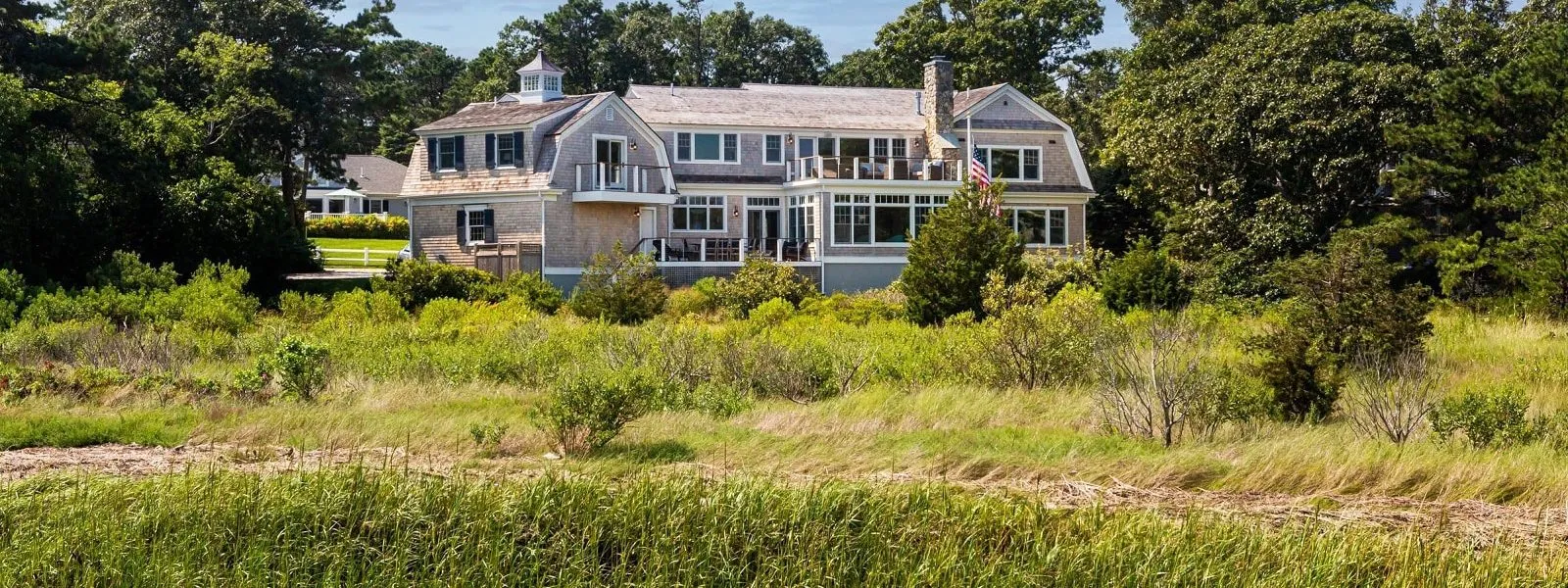 A two-story wood-sided house amid trees and tall grass by water – McPhee Associates of Cape Cod.