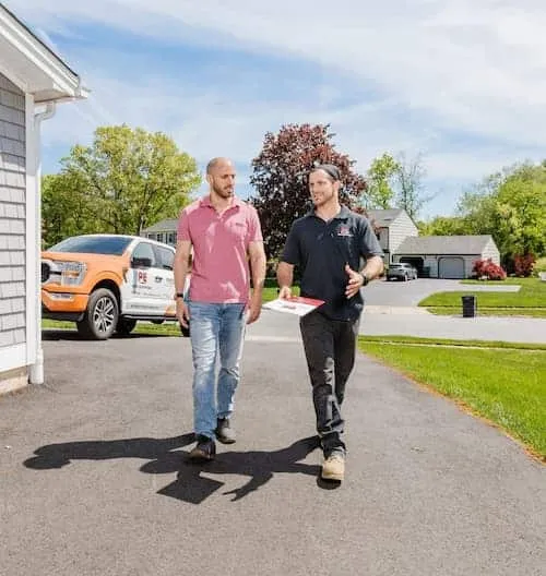 Two men approach a home, one in a PE Home Generators shirt holding a clipboard.