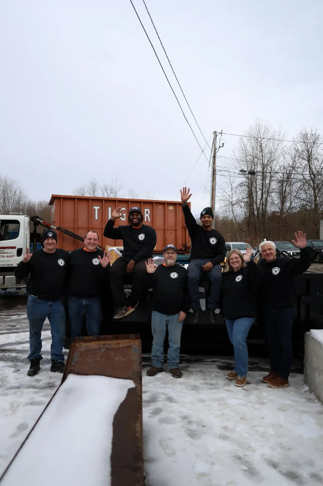 Group of seven people standing outdoors on snowy ground, waving at the camera, with a truck and trees in the background.