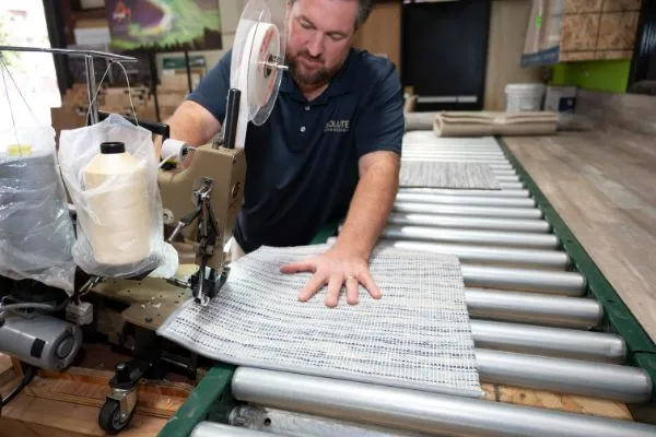 A man sews carpet on a conveyor belt, likely during carpet and floor installation. The sewing machine is large and industrial. The man is focused on his work.