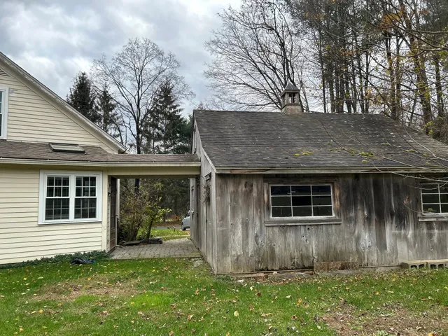 A rustic wooden shed next to a white house under a cloudy sky.