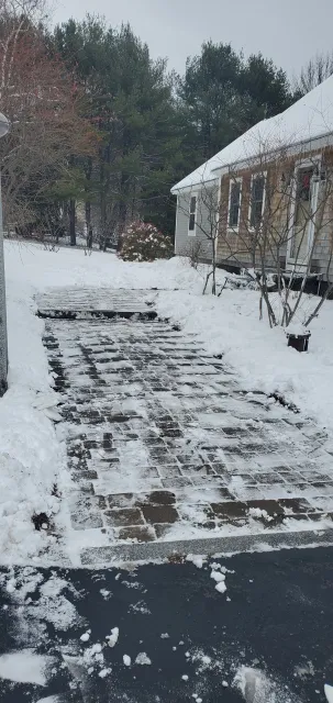 Snow-covered driveway with cleared pathway, flanked by trees and a house.