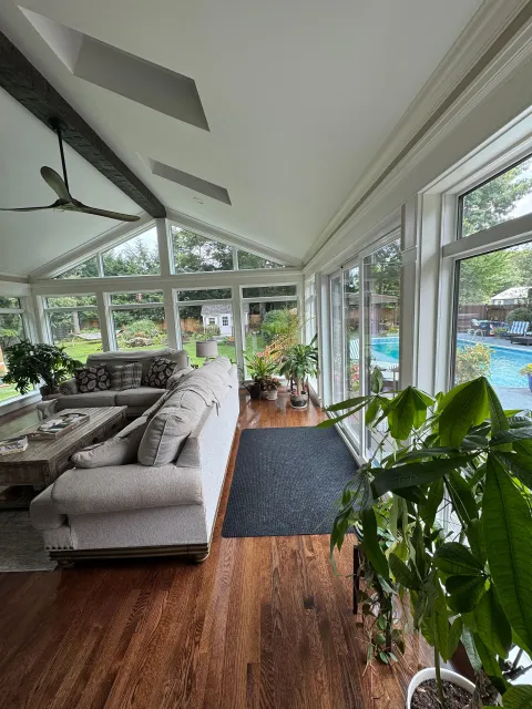Bright sunroom with gray sectional sofa, wood floor, indoor plants, and large windows overlooking a pool.
