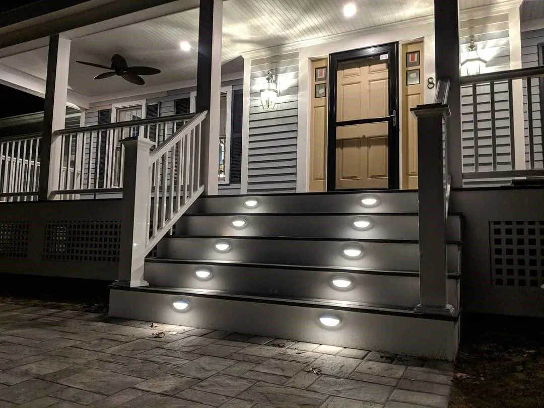 Nighttime view of a front porch with stairs leading up to a door, illuminated by inset porch lights, with a ceiling fan and exterior lighting on the house.