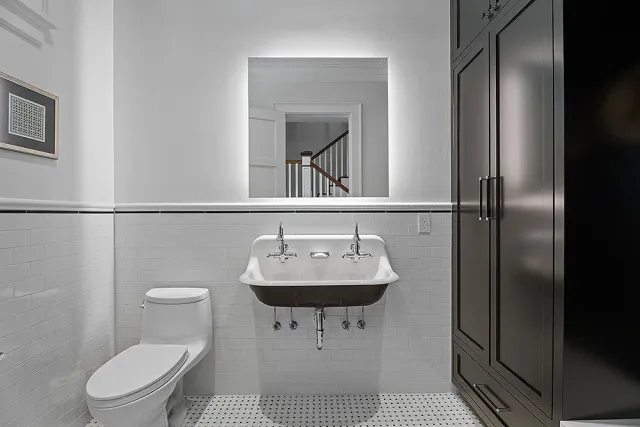 A modern bathroom with white tile, toilet, sink, and dark cabinet. A mirror is centered above the sink.