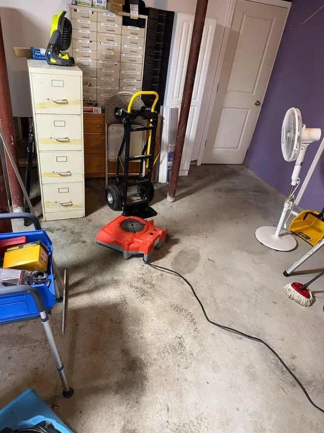 Garage cluttered with cleaning tools, a red floor machine, filing cabinet, and a standing fan on concrete floor.