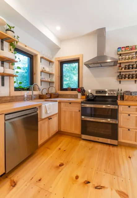 Kitchen with wooden cabinets, stainless steel stove, open shelves with jars and cups, windows with greenery outside, wood flooring, and a range hood.