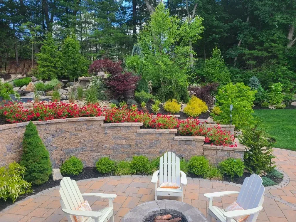 Three Adirondack chairs sit on a stone patio around a circular fire pit in front of a tiered, garden-filled retaining wall.