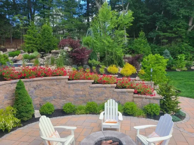 Three Adirondack chairs sit on a stone patio around a circular fire pit in front of a tiered, garden-filled retaining wall.