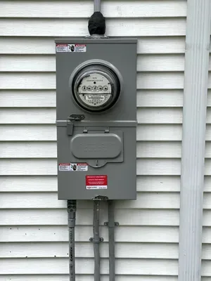 Gray electric meter box mounted on white siding with conduit pipes below