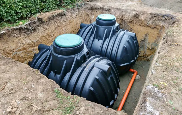 Three dark septic tanks installed in a dirt excavation, connected by orange pipes, and ready for backfill.