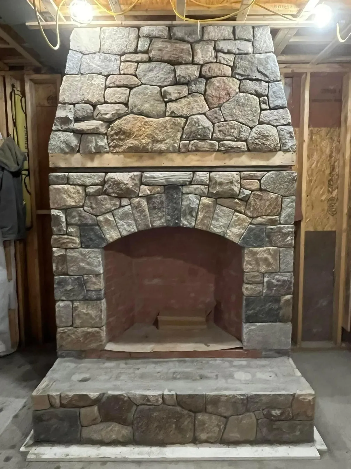 A constructed stone fireplace with a curved arched opening, built with various sizes of gray and brown stones, in an unfinished basement with visible wooden framing and wiring.
