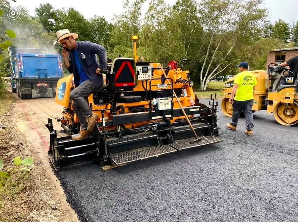 A construction worker uses a phone while operating yellow asphalt paving equipment on a road next to a roller and dump truck.