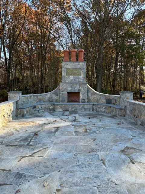 Stone outdoor fireplace with chimney pipes, surrounded by a stone patio and wooded area in the background.