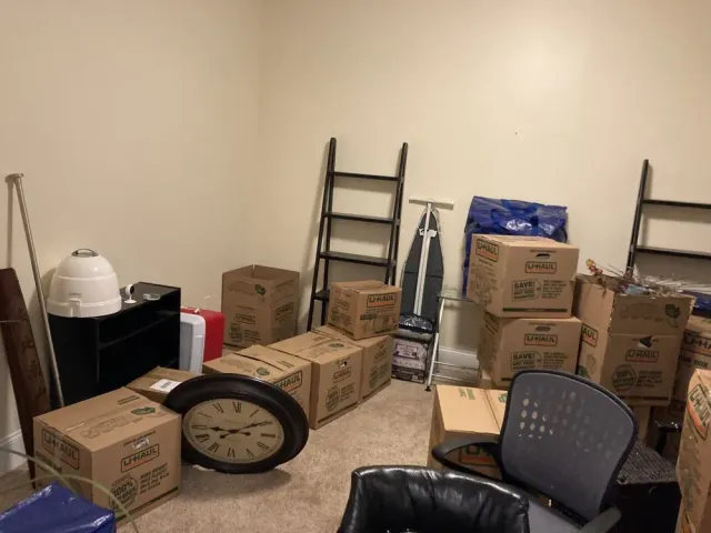Room filled with cardboard boxes, a clock, ladder, and ironing board, indicating a move or storage.