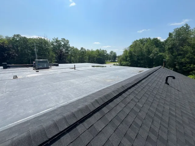 View of a flat commercial roof with rooftop equipment beside a shingled sloped roof, surrounded by trees under a clear blue sky.
