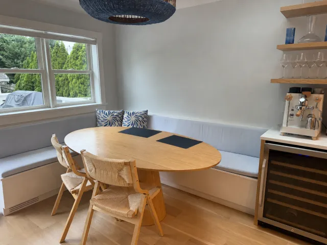 Dining nook with a round wooden table, a built-in white bench with cushions, two chairs, large window with a view of green trees, a beverage station with wine glasses, a coffee machine, and a wine cooler.