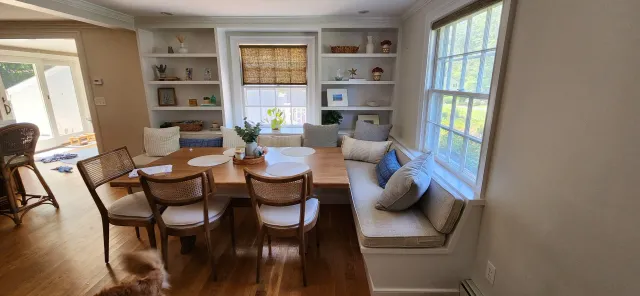 Cozy dining area with wooden table, chairs, and built-in bench seating. Shelves with decor items flank a large window, and a small dog is visible on the floor.