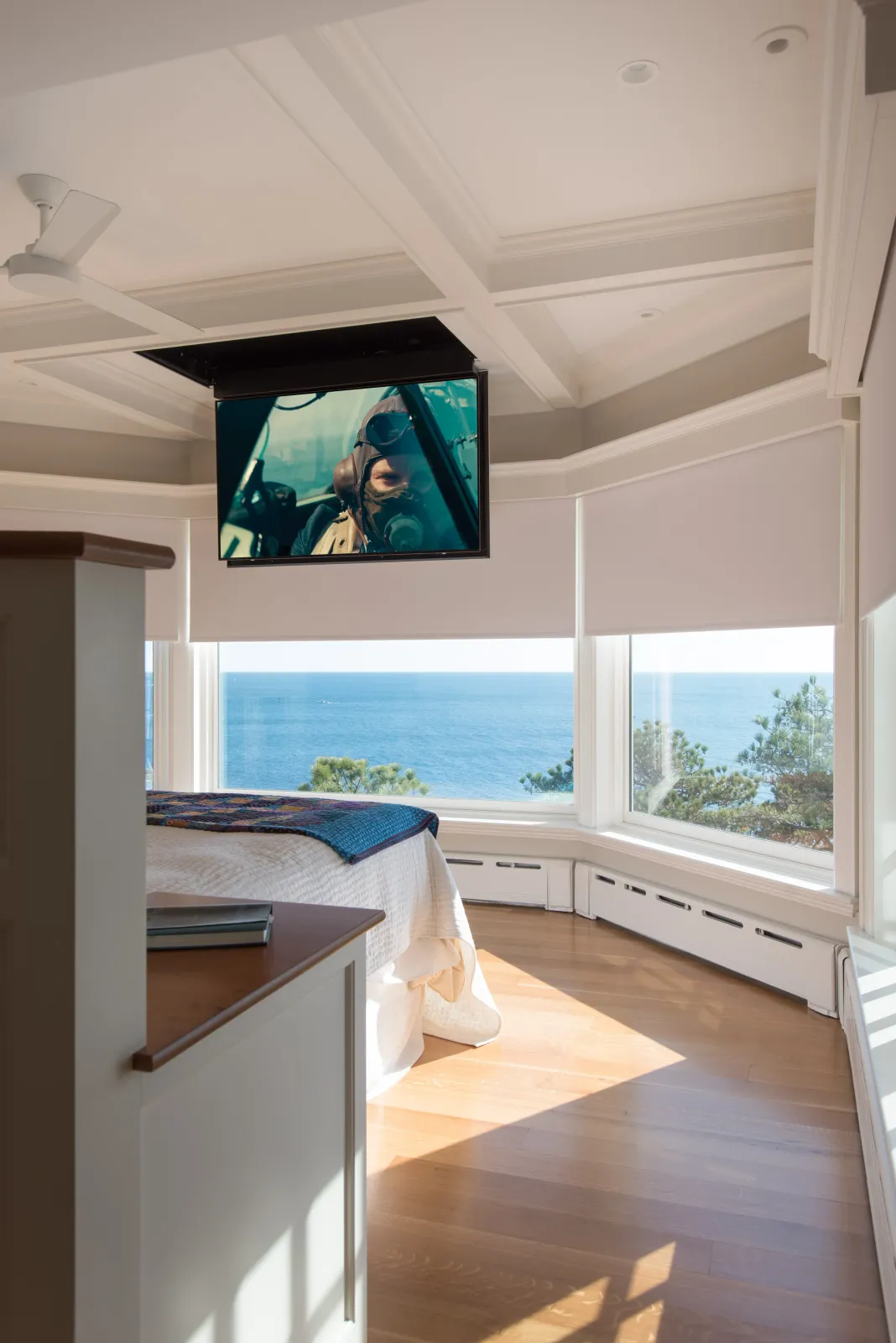 A bedroom with large windows overlooking the ocean, a bed, a blanket, a TV mounted on the ceiling, and sunlight on the wooden floor.