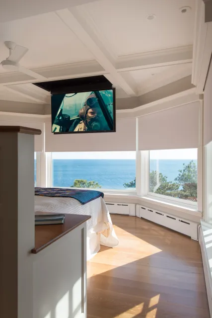 A bedroom with large windows overlooking the ocean, a bed, a blanket, a TV mounted on the ceiling, and sunlight on the wooden floor.