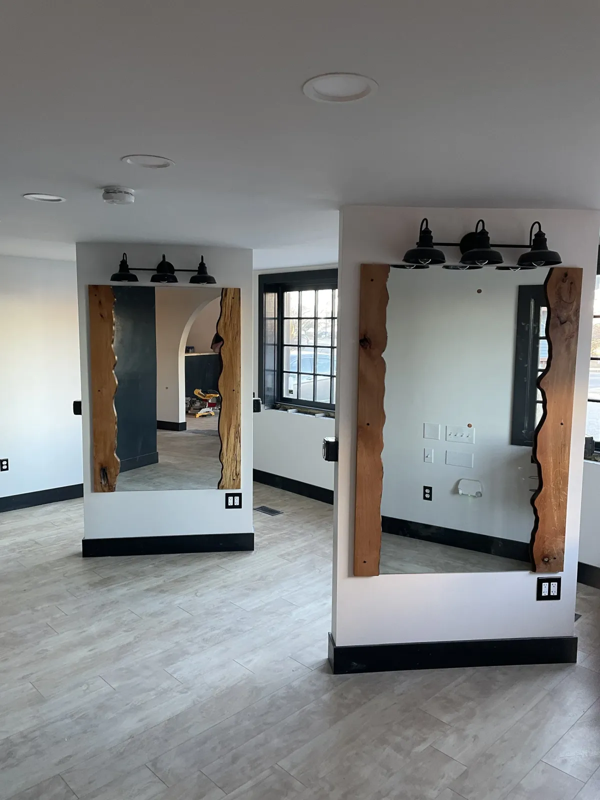 Interior space with two mirrors framed with natural wood, black light fixtures above, a window with black framing, and wood flooring.