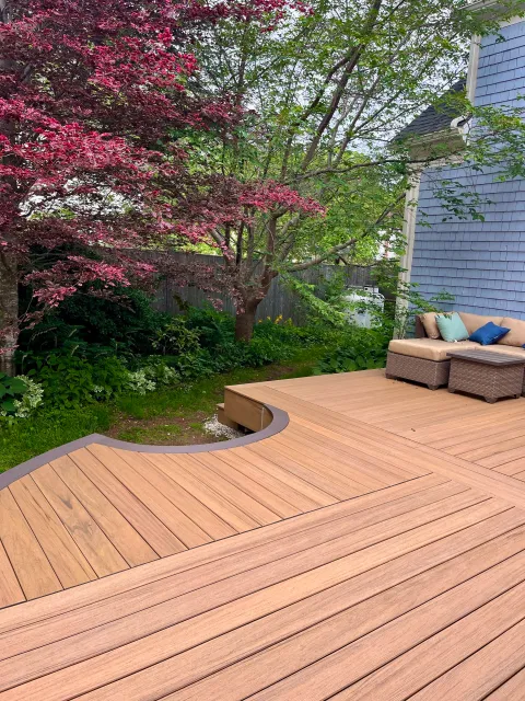 Outdoor backyard scene with a wooden deck, a table with cushions, greenery, and a house with blue siding.
