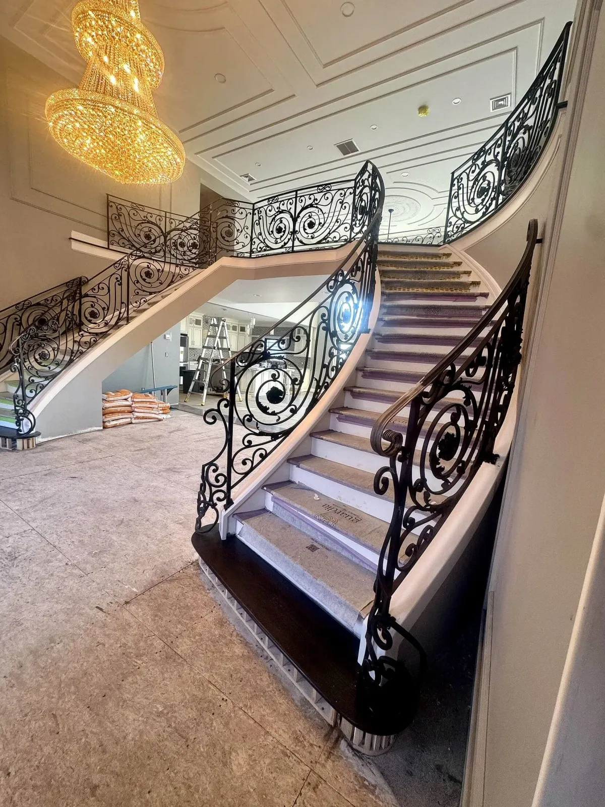 An indoor staircase with ornate black wrought iron railings and beige steps, with a large sparkling gold chandelier hanging from the ceiling, in a well-lit luxurious interior space under construction or renovation.