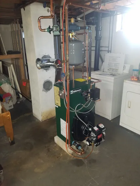 Green boiler with copper pipes in a basement next to a washing machine.