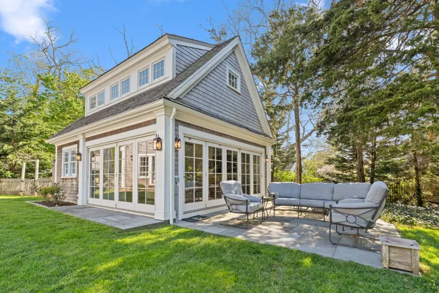 Exterior back of a custom Cape Cod home with a stone patio addition, French doors, gray wood shingles, outdoor seating area, and a well-maintained lawn