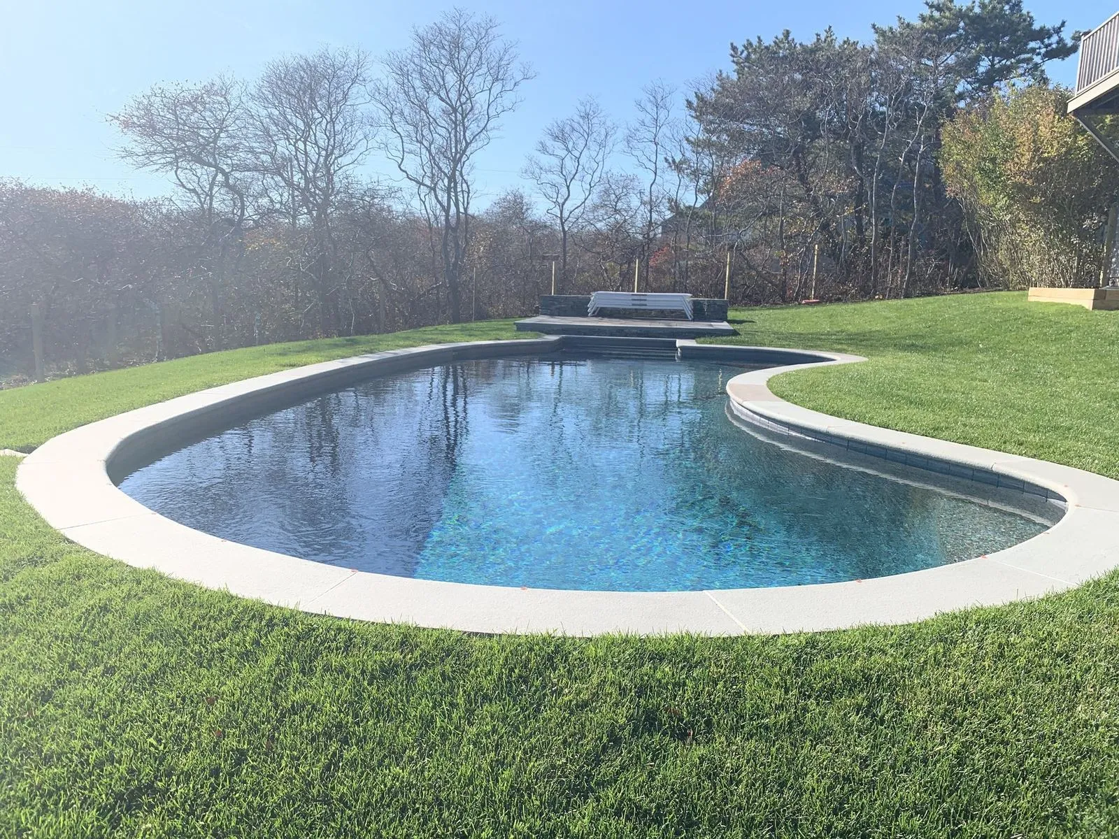 A curved free-form pool with a stone-clad spa in a Cape Cod backyard, framed by lawn and a coastal tree line